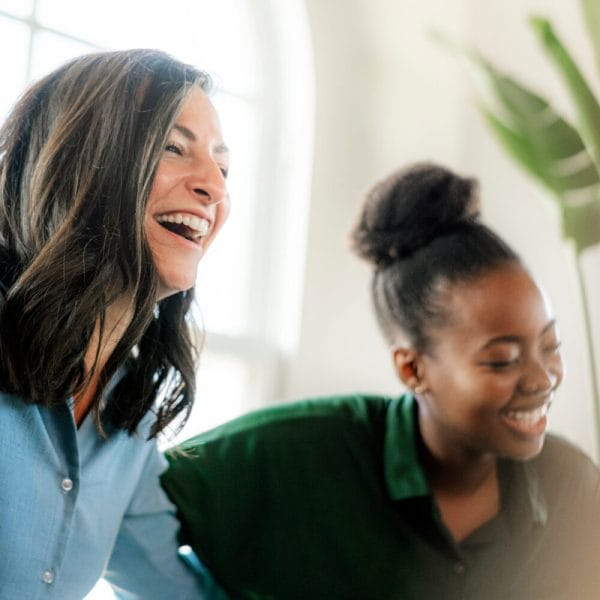 A white woman with short brown hair and a young black woman with her hair in a bun are laughing in a meeting.