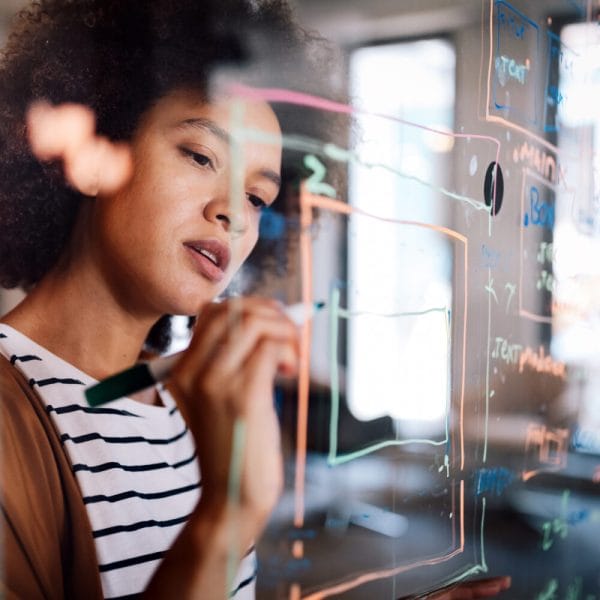 A young woman with an Afro and light brown skin is drawing a diagram and making notes on a glass board in an office.