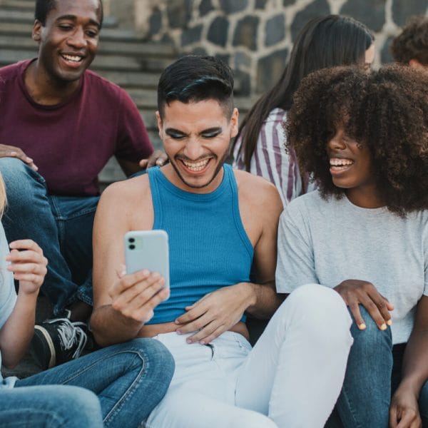 A diverse group of people having fun. A gender non-conforming person holds a cell phone, while laughing with a Black woman with an Afro and a black man.