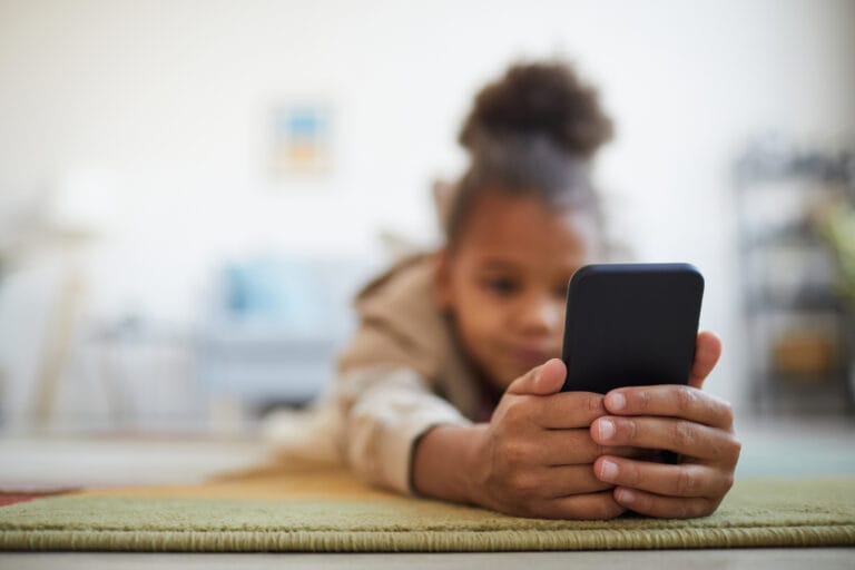 Close up of at cute African-American girl holding smartphone while lying on floor in cozy home interior